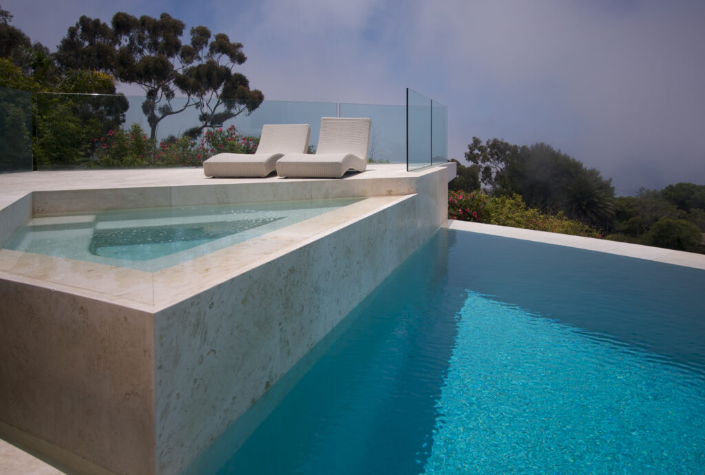 A high-end hillside pool with a marble finish, an elevated hot tub surrounded by a glass protective wall, and two lounge chairs, designed by Bakersfield Top Pool Builders.